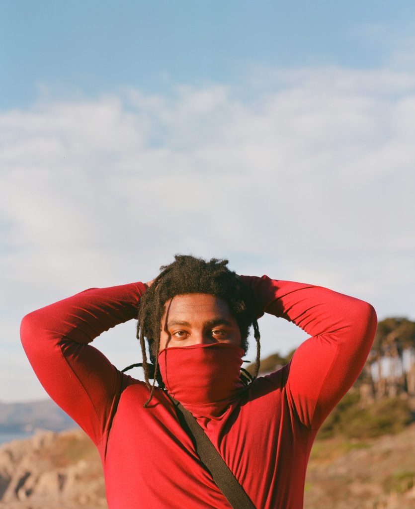 Lil B looking straight into the camera. The blue sky and clouds make up 2/3 of the frame at the top. He fills up the bottom 1/3 of the frame wearing a red turtle neck with his arms bent at the elbows and resting behind his head. The red neck of his shirt is pulled up to cover half of his face. He is wearing his hair in loose dreads.