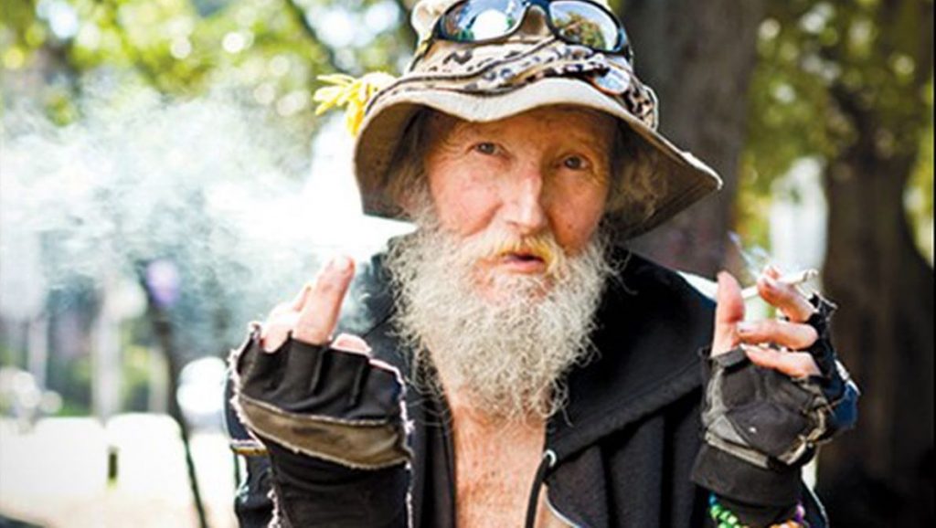 Hate man stares into the camera, flipping the photographer with his right hand and holding a lit cigarette in his left. He wears a bucket hat on his head with a bandana wrapped around it, several pins, and a flower, with sunglasses perched on top.