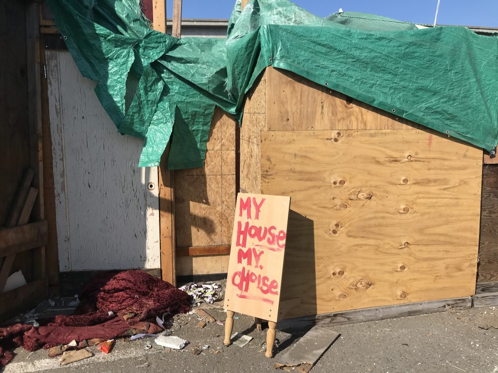 A self-built wooden structure covered with a green tarp. In front a sign reads "my house my choice," although the word "choice" is misspelled--it is spelled "c-h-o-i-s-e."