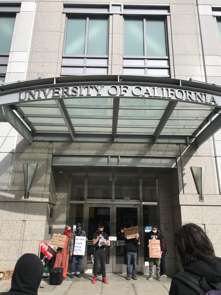 A group of young people hold signs that read "no-one owns mama earth," and "Stop sweeping us" underneath the OC office of the chancellor building. Above them is a big sign that sys "University of California"
