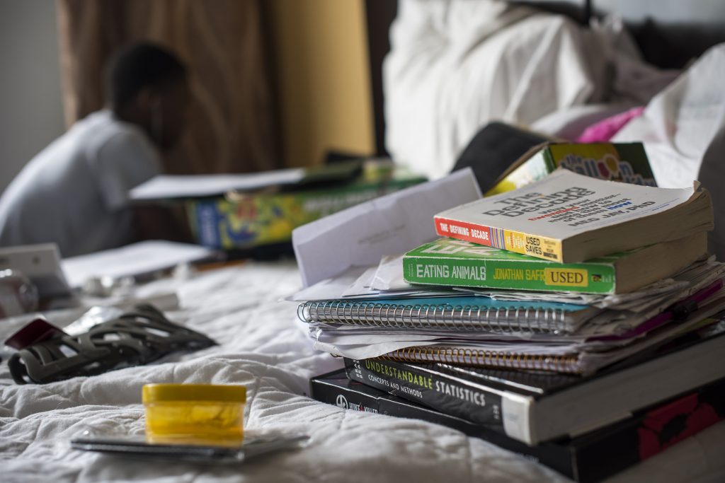 A hotel ned with a pile of books and notebooks on it. In the background, the silhouette of a man wearing headphones is blurry but visible.