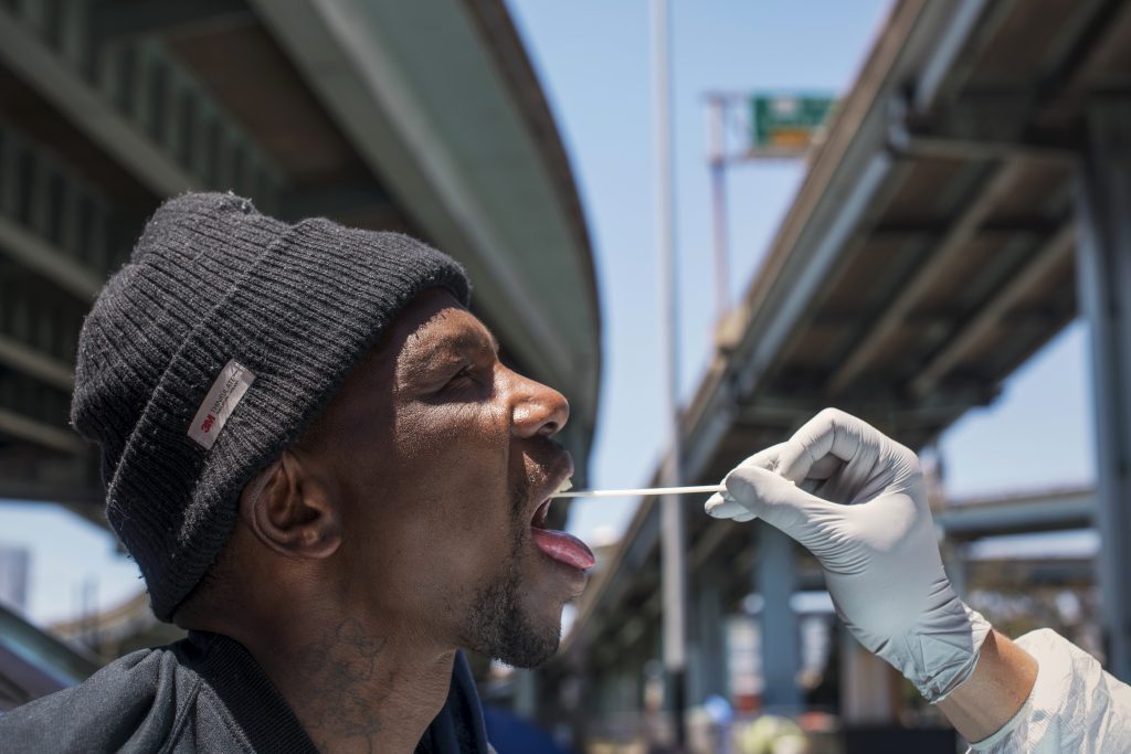 A man in a beanie opens his mouth and a white gloved hand inserts a long cotton swab into his mouth to test him for the coronavirus. His face is framed by the highway overpasses above him--two concrete paths with blue sky shining between them.