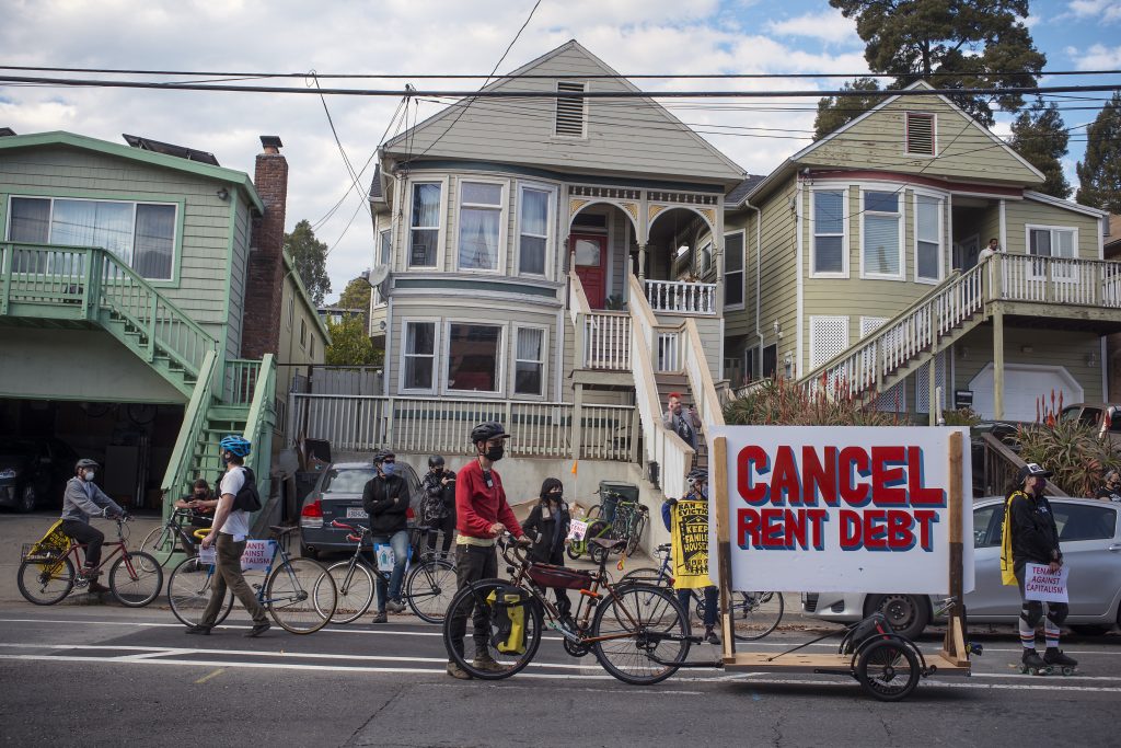 Protestors on bicycles stand on the street with a sign that says "cancel rent debt" in front of three victorian houses in Oakland.