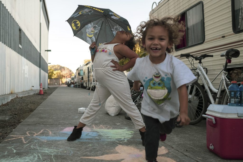 Two children dance and play on the street outside of their RV. Multicolored chalk drawings can be seen on the sidewalk beneath them.