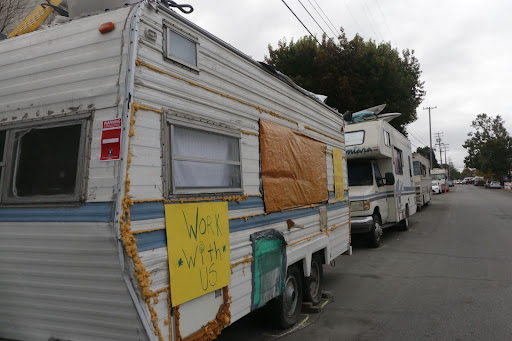 A line of RVs along Eighth street. One of them has a sign taped to the side reading "work with us."