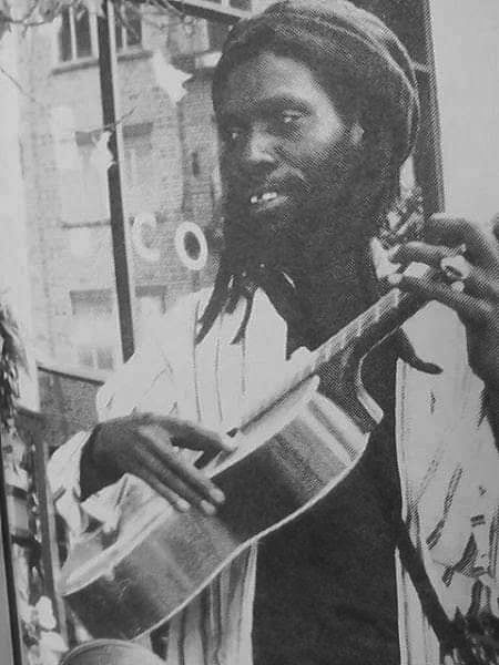 A black and white image of Bledsoe, a Black man with dreadlocks playing an acoustic guitar. 