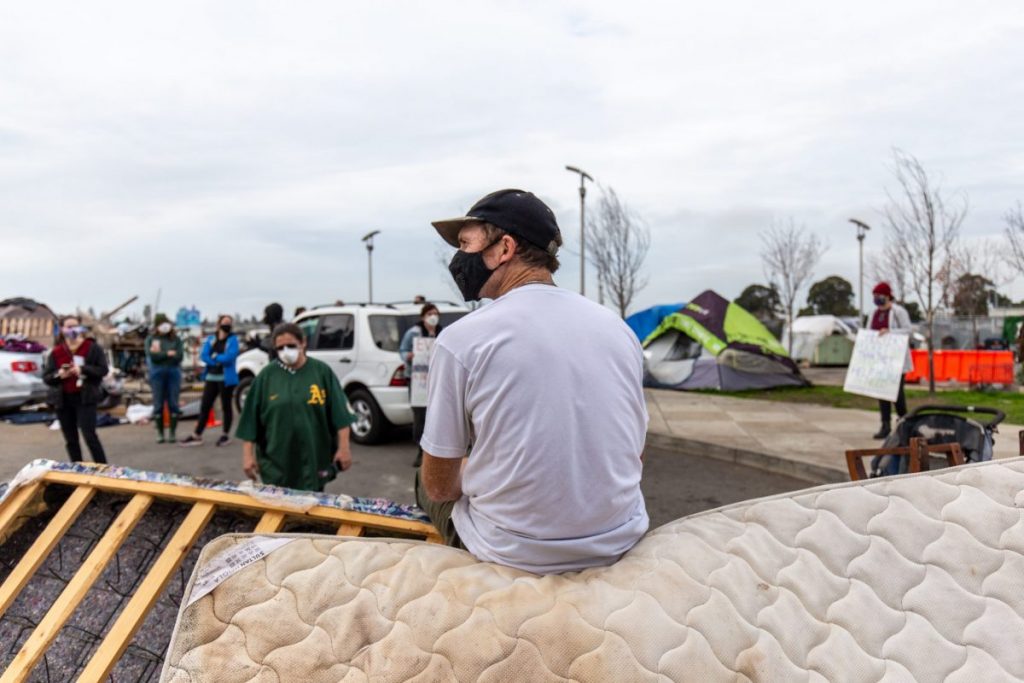 An encampment resident sits on a barricade made out of a mattress with his back to the camera. Behind him, protestors and tents can be seen, facing the camera.