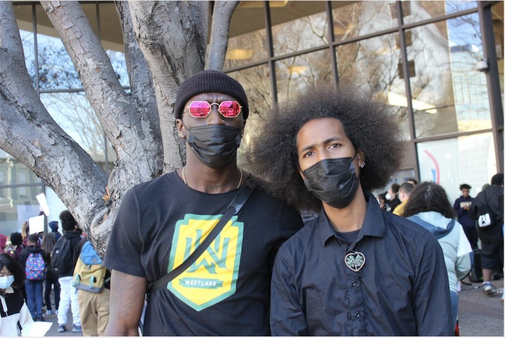 Moses and Andre, both Black men, stand outside a government office, wearing black face surgical masks and looking intensely into the camera.