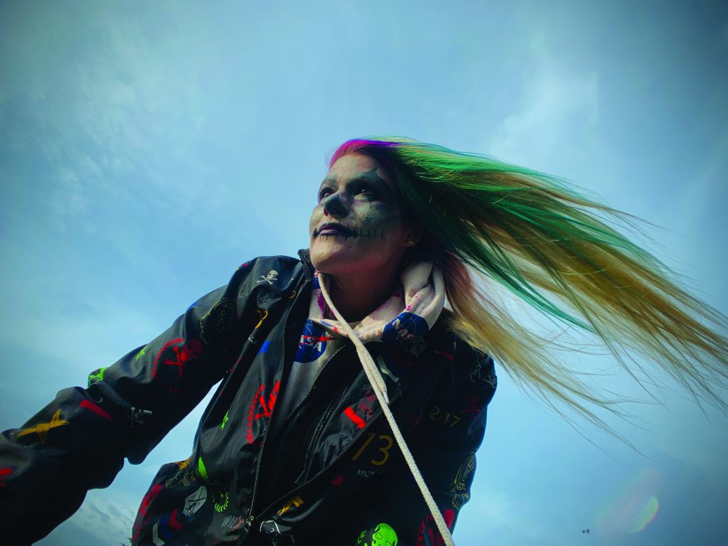 Ashley, a white woman who looks to be in her 20s or 30s, wears rainbow paint in her face and hair. The photo is taken from below. She looks to the side and smiles white her hair blows to the side. The sky is blue behind her head. 