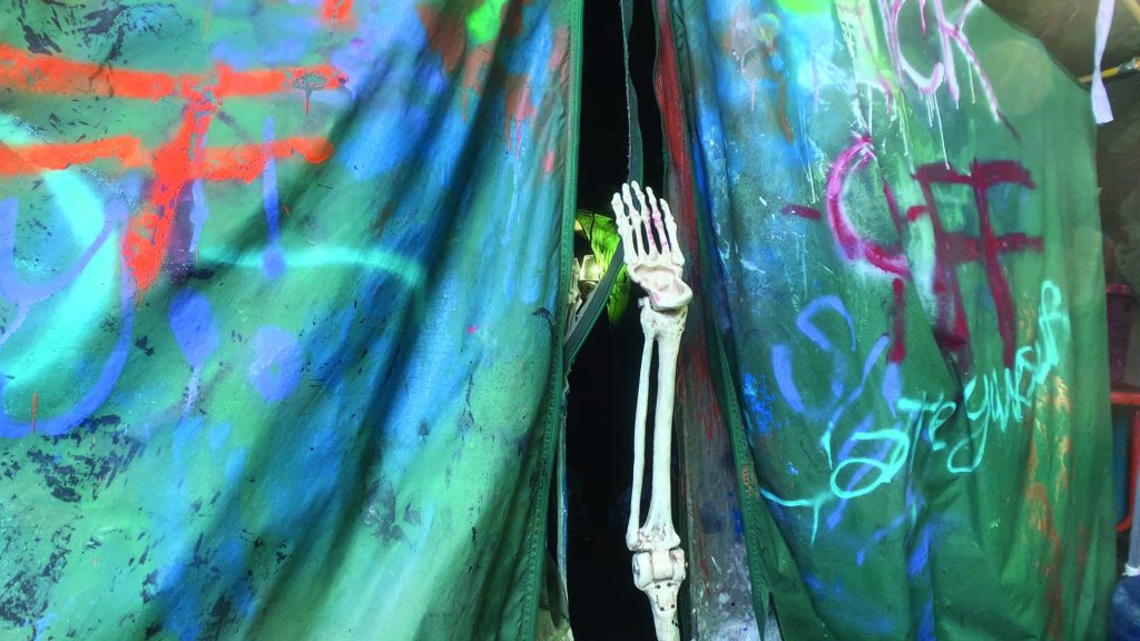 The side of what looks like a teal colored tent with multi-colored graffiti scrawled all around it. In the center, there is an opening to the tent. The opening frames the white hand of a skeleton, which reaches up from below.