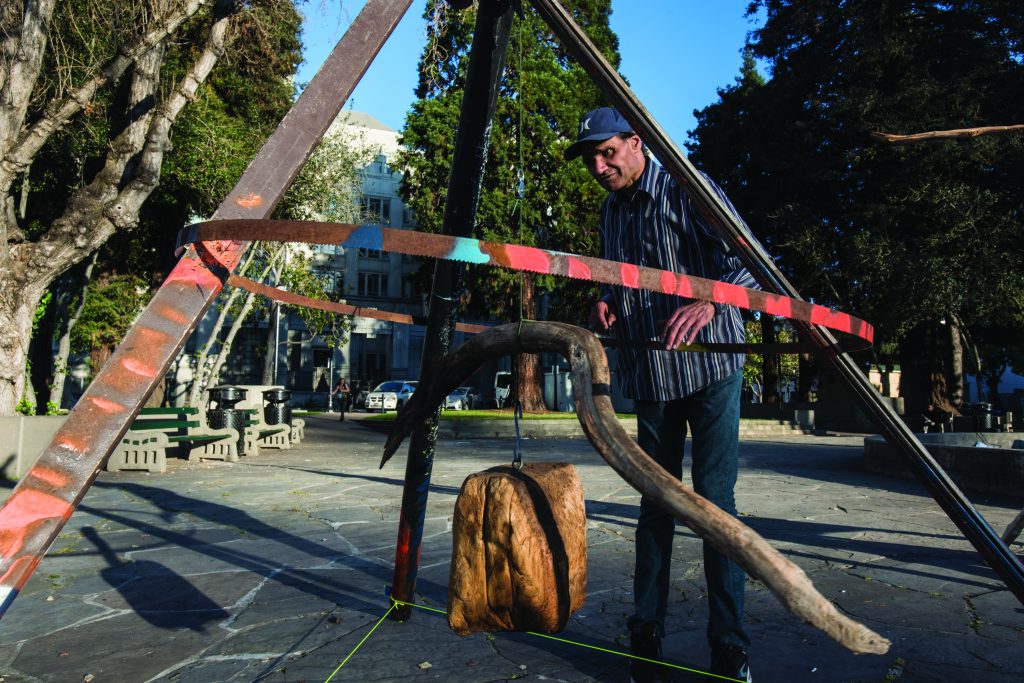 D'art Lloyd sets up one of his sculptures at MLK Civic Center Park. The sculpture is comprised of three metal poles that are arranged in the shape of a pyramid, with a circular metal hoop around them. All the bars plus the hoop are painted, stripes of red and blue. In the middle of the pyramid shape a two large pieces of driftwood hang from a string.