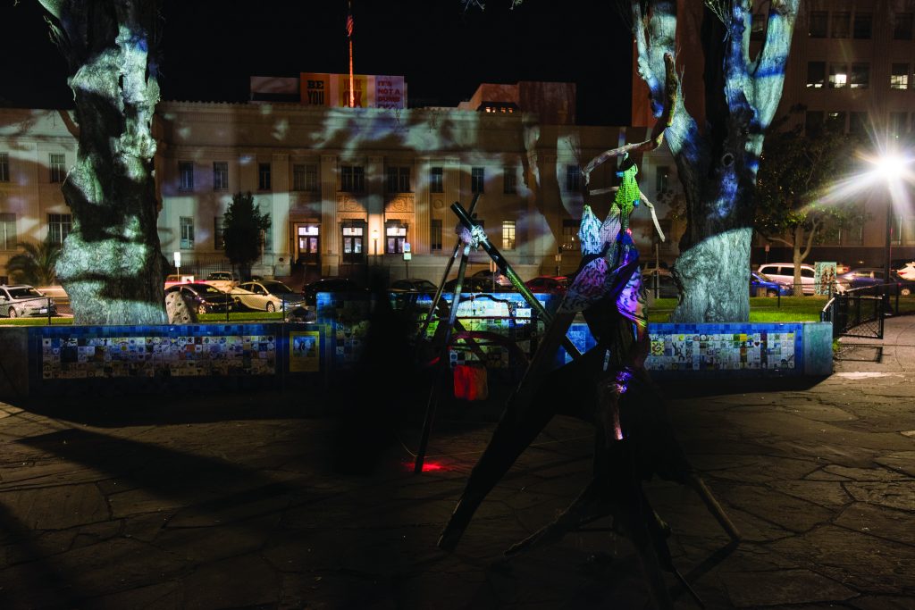 An image of Civic Center park at night time. In the foreground there are two sculptures in the shape of teepees. In the background, images are projected onto trees and buildings. The images are not crisp and clear but rather more abstract, giving the scene the feeling of being under water.