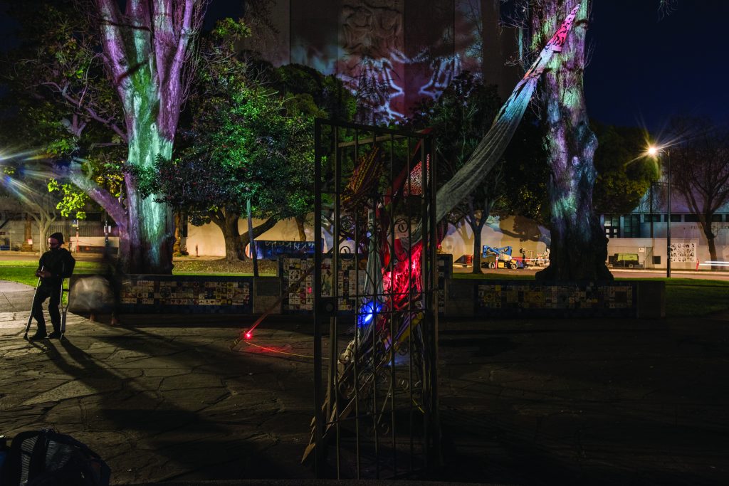 An image of Civic Center park at night time. In the foreground there is a sculpture made out of a metal door. In the background, images are projected onto trees and buildings. The images are not crisp and clear but rather more abstract, giving the scene the feeling of being under water.