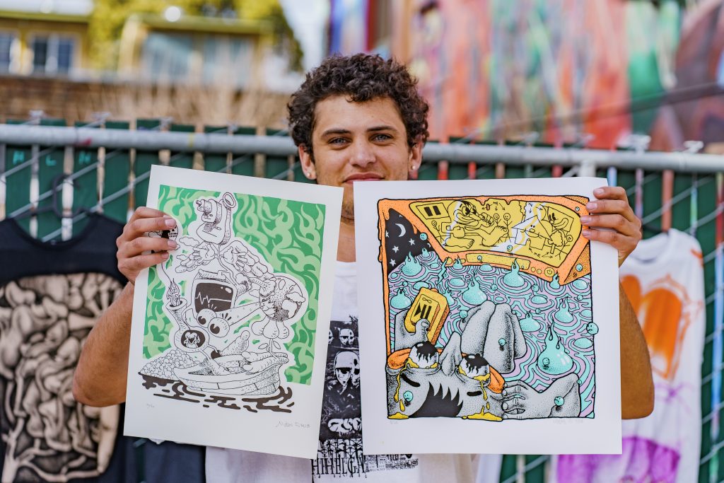 An artist holds up two drawings that they made, one with dominant green colors and the other with an orange-based color pallet.