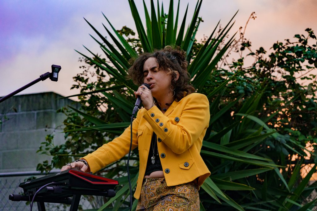 A musician sings into a microphone and plays the keyboard. The sun sets behind them and they are wearing a yellow jacket.