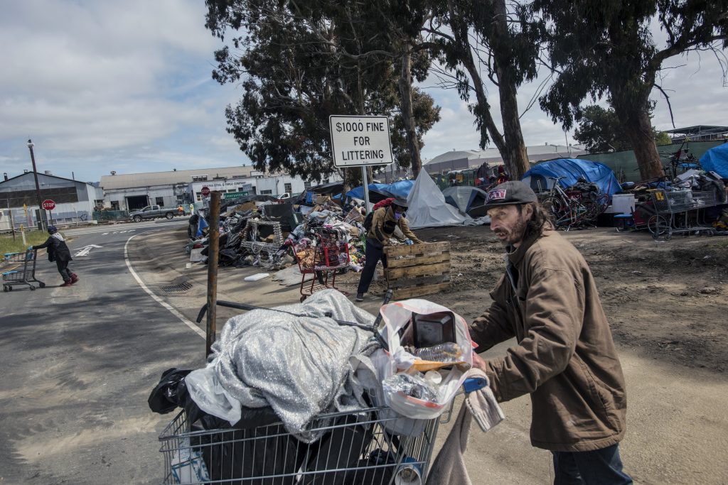 An unhoused man pushes a shopping cart full of materials across a freeway offramp. The rest of the encampment surrounds a sign that reads "$1,000 fine for littering" in the background.