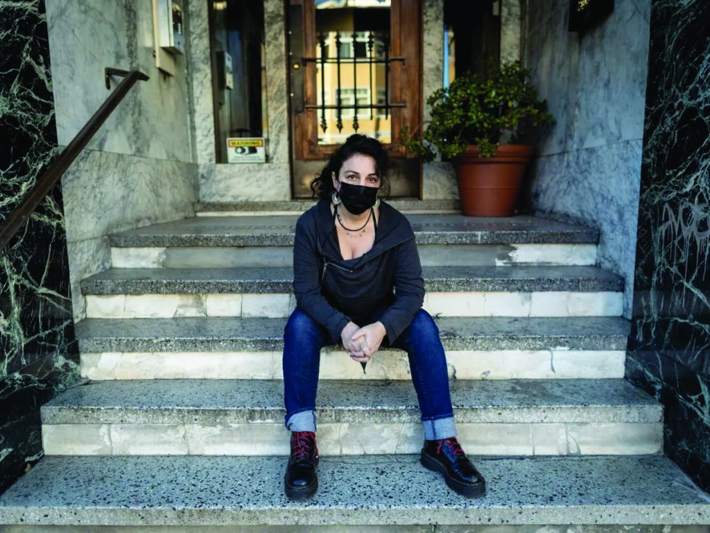 Christina sits on the steps of her building wearing a mask.