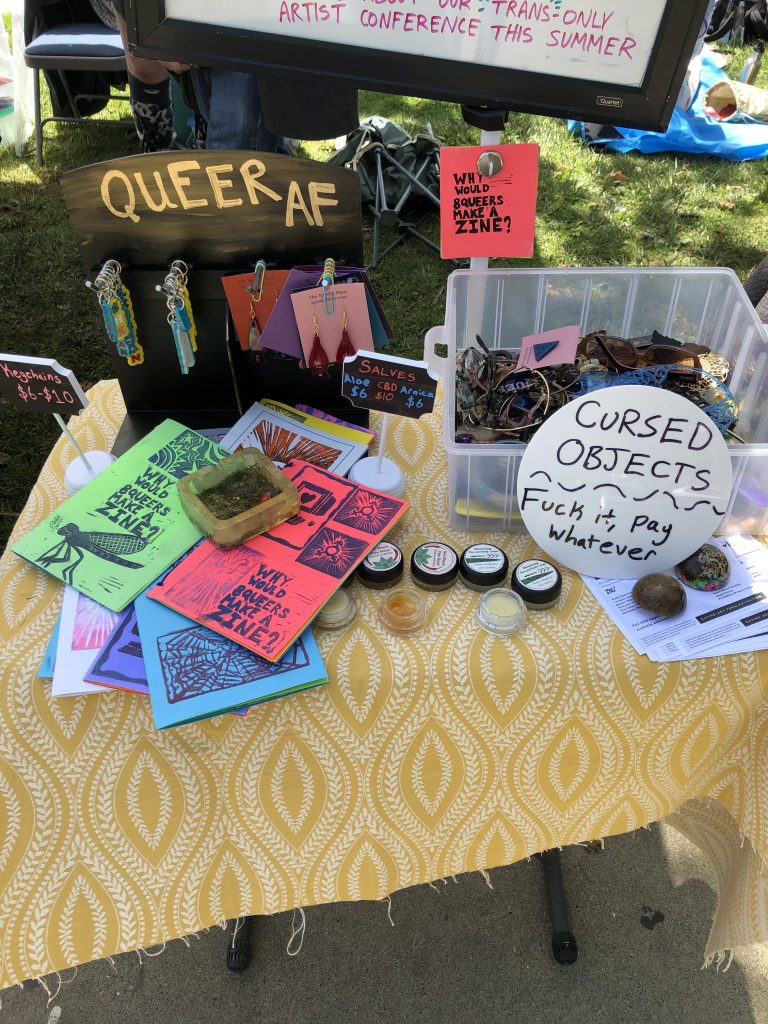 A table full of wares being sold at the art market, such as zines, keychains, and a bucket labeled "cursed objects." A sign reads "Queer AF."