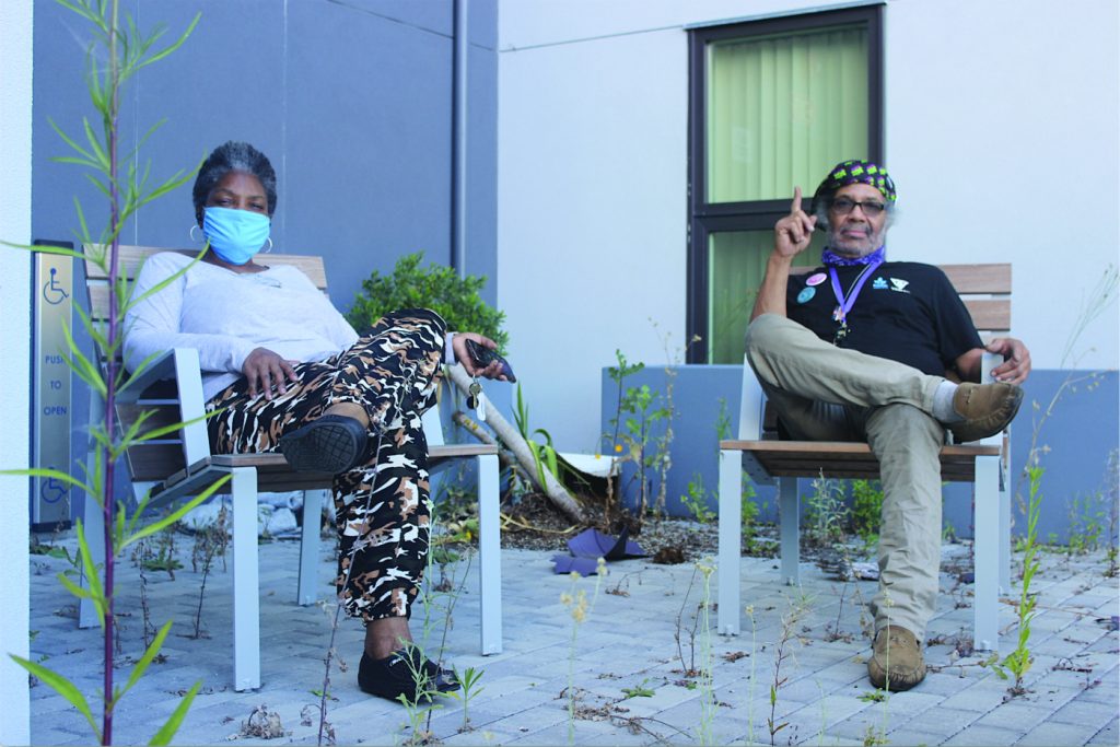 Two Black tenants of Embark sit in an in outdoor furniture, looking at the camera. Around them, tall weeds sprout up from the tiled patio floor.