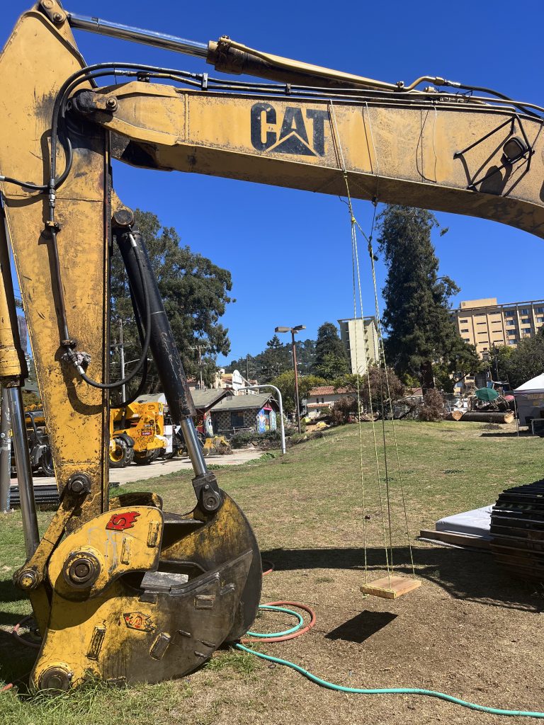 A large yellow CAT excavator has a wooden swing tied to the scooping arm.