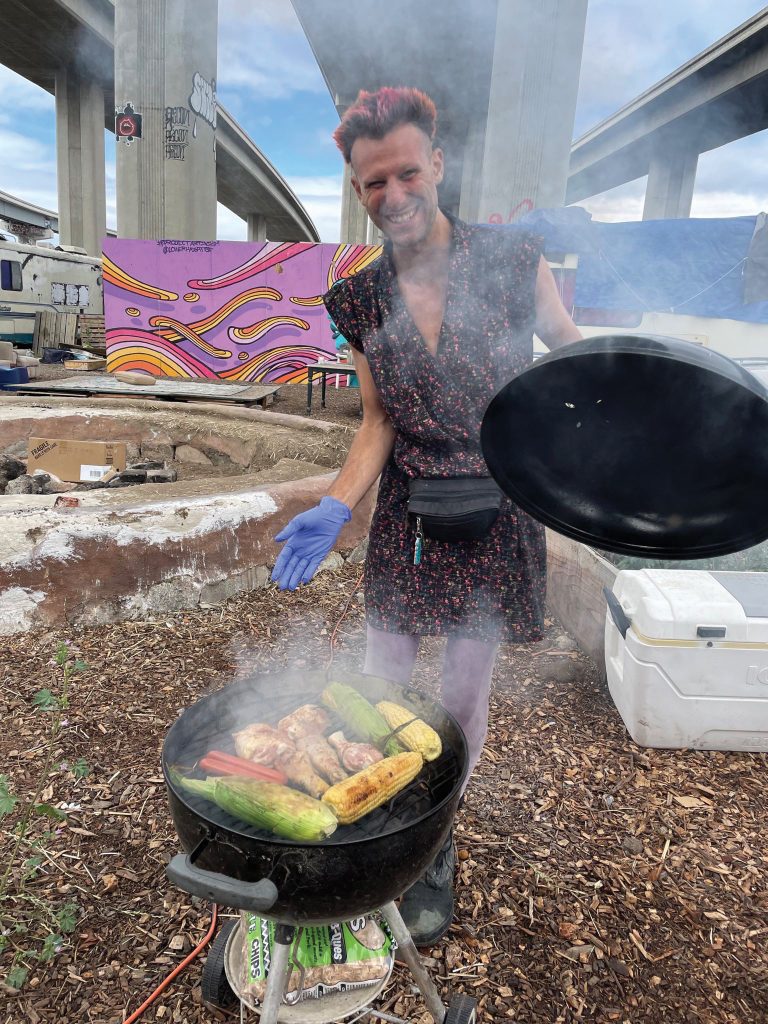 Wood Street resident Jaz Colibri has pink hear and wears a floral dress. She is barbecuing chicken and corn on a small BBQ at the encampment.