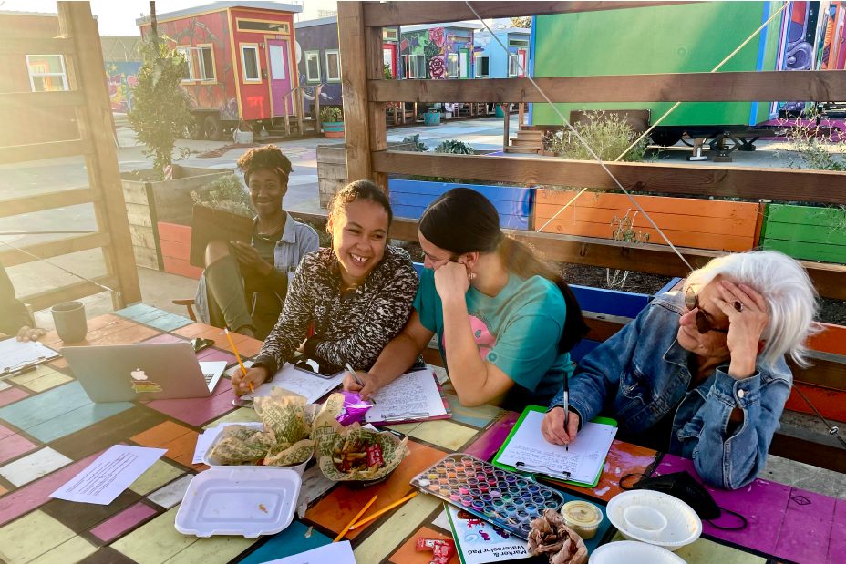 The writer's workshop takes place on a colorful picnic table. Participants laugh together with paper in front of them and pencils in their hands.