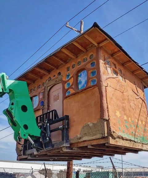 A cob home—a tiny house that looks as if it was made out of mud—hovers in the air as it is lifted up by a green forklift in front of a blue sky.