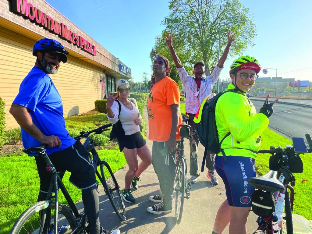 A group of unhoused people and their advocates wearing neon yellow and orange pose for a photo in front of their bikes.
