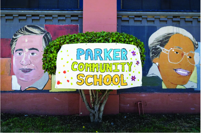 A mural on the side of the school in the background. In the foreground at tree with a colorful sign pinned on it reading "Parker Community School."
