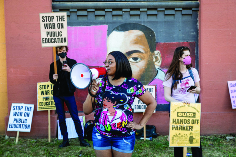 Three protestors stand in front of a mural Martin Luther King Jr. The middle protestor is a black woman speaking into a microphone. In the background are two white protestors in masks holding signs that say "stop the war on public education," and "OUSD: hands off Parker!"