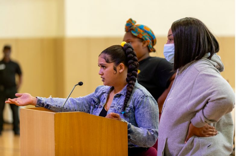 A group of three women speak at a podium at a school board meeting.