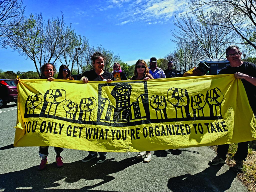 A group of white people hold a large, long yellow banner depicting many solidarity fists above the words "you only get what you're organized to take."