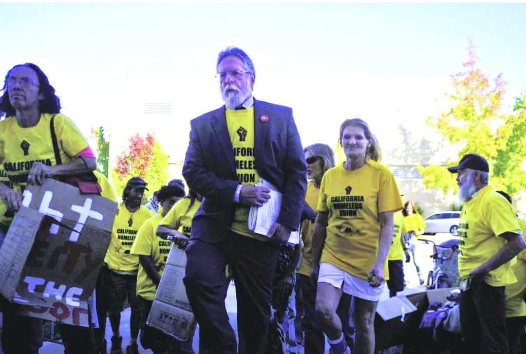 Anthony Prince, a white man, walks in a group of people wearing a yellow shirt that says "California Homeless Union"