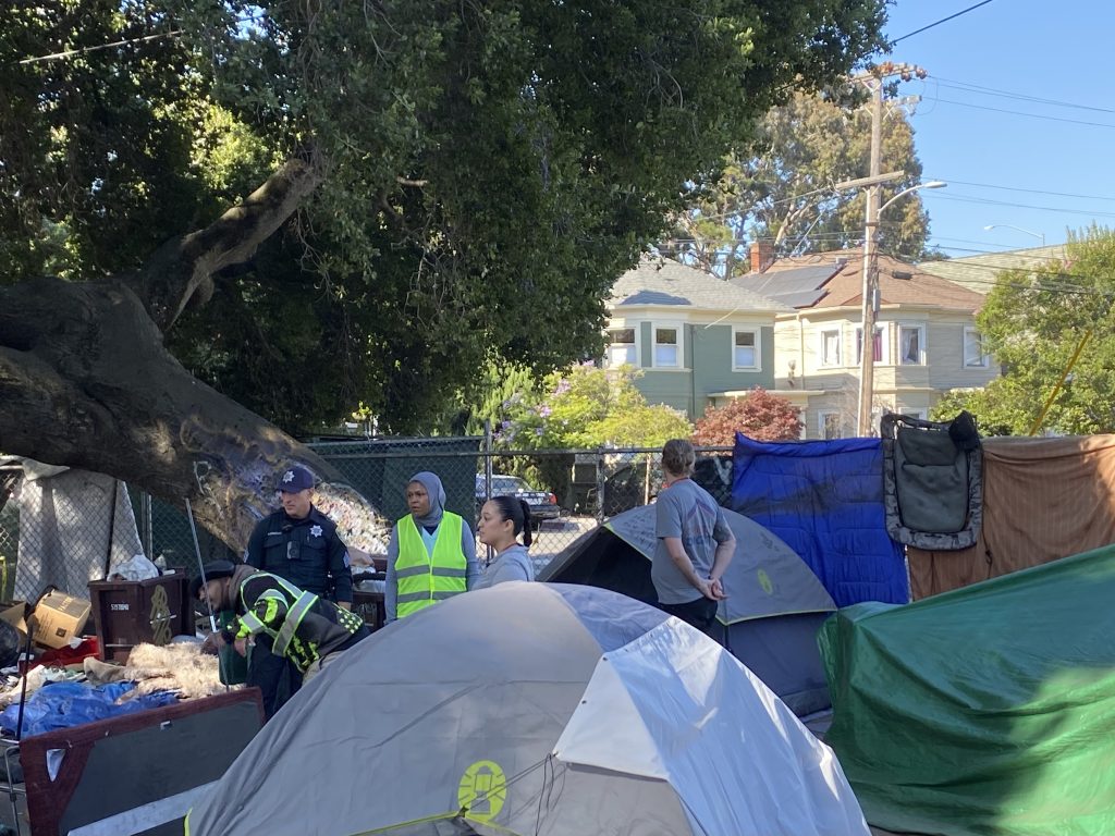 Multiple tents are huddled against the Webster Street fence line in Mosswood Park. Members of Oakland's Encampment Management Team are speaking to a resident outside of their tent, including representatives from the City Administrator's office, Oakland Police Department, and Operation Dignity.