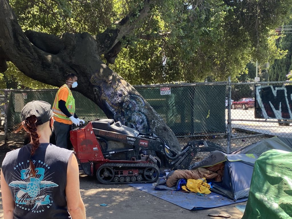 An employee of Oakland's Department of Public Works removes a tent with a track loader as onlookers watch in the foreground.