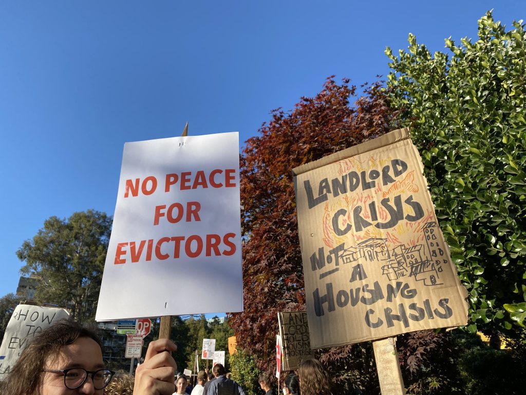 Two picket signs are held by tenants protesting the event, reading "No peace for evictors" and "Landlord crisis, not a housing crisis."