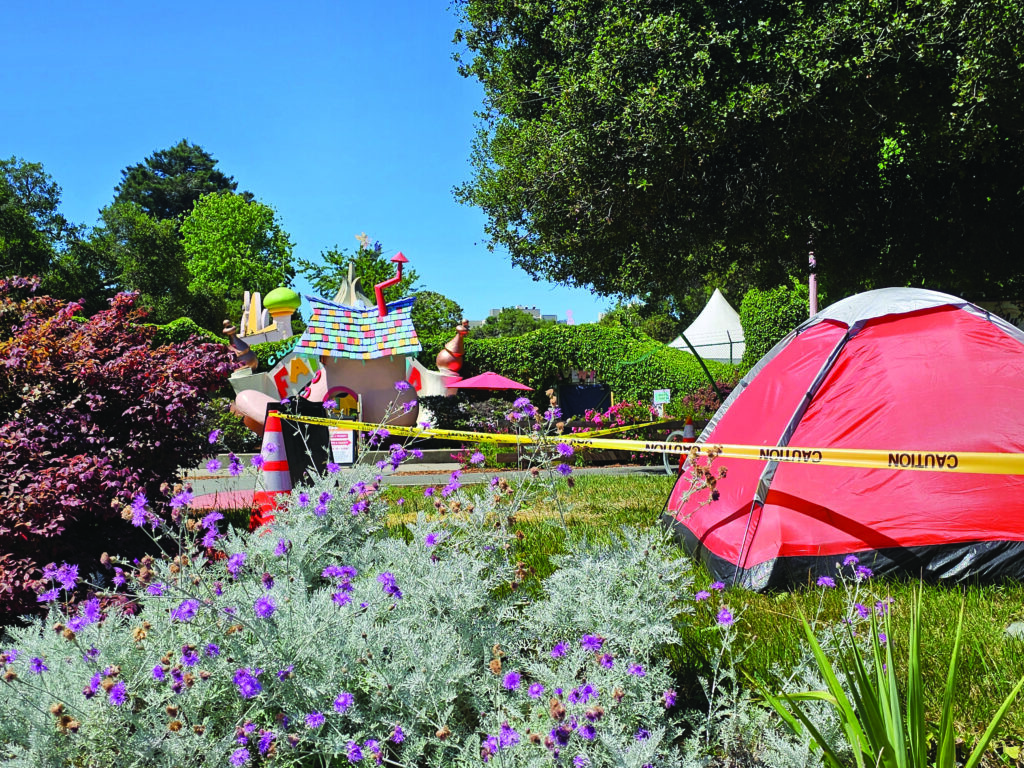 Photograph of a children's storybook theme park and the red tent of an encampment resident in front of it. The tent has caution tape tied around it so that no one but city workers can come near the tent but city workers participating in the encampment sweep of this area.