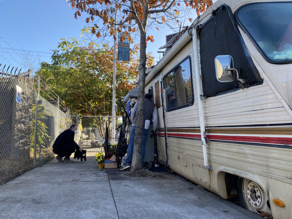 A white man peeks into the window of an RV, where somebody lives. Another white man wearing a mask crouches down and pets a black cat in front of the RV.