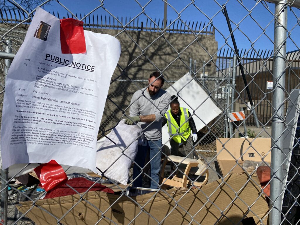 Through a chain link fence, the viewer can see city officials work on packing up the possessions of encampment residents and preparing them for removal. There is a "public notice" taped onto the fence, instructing encampment residents to vacate the area.