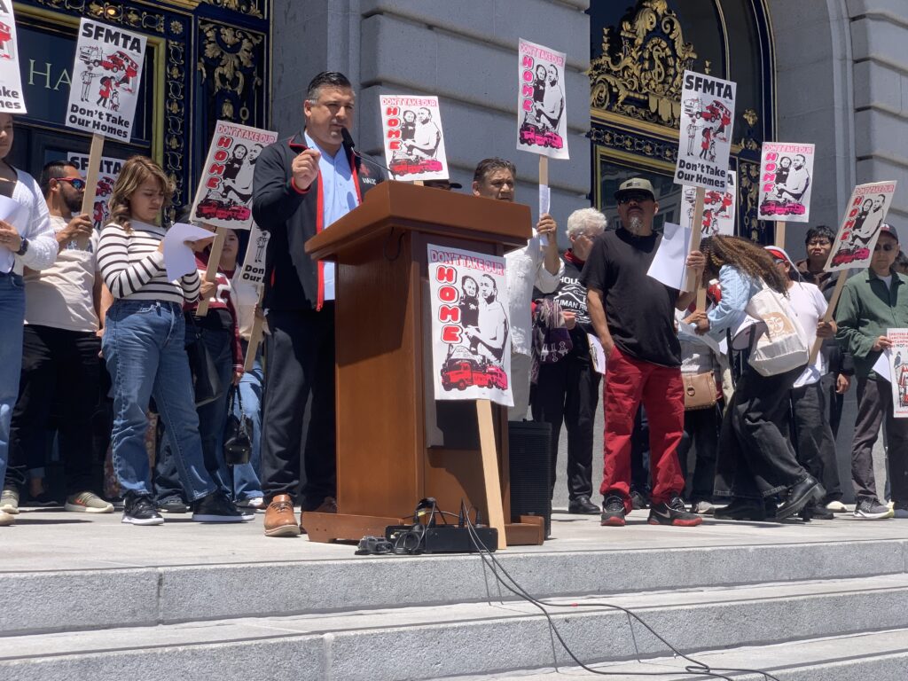 A Latinx man in a black and red jacket stands at a podium in front of San Francisco's City Hall and speaks into a microphone. All around him protestors carry signs that say "don't take our homes"