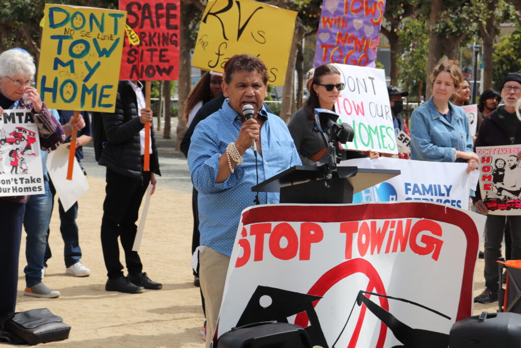 A Latinx man in a blue shirt stands at a podium in and speaks into a microphone. All around him protestors carry signs that say "don't tow my home" and "stop towing."
