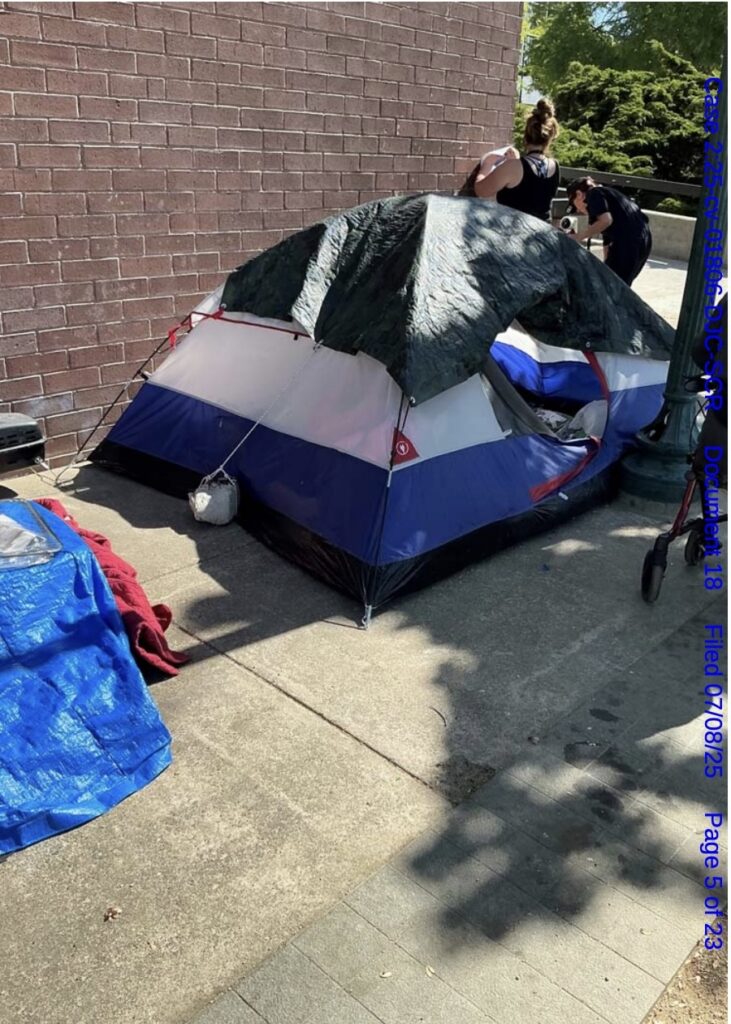 Two people can be seen packing up behind a blue and red tent, with a black tarp over the top. They are facing away from the camera. The tent is sitting in front of a brick wall.