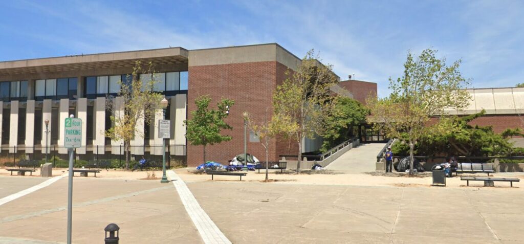 A street-view image of the John F. Kennedy Library, which is a brick building with a few trees out front. A few tents are set up against the front of the library.
